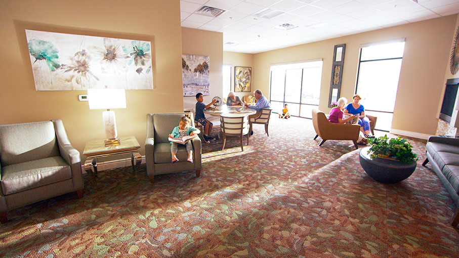 A spacious lounge area with several people seated and interacting, filled with natural light from large windows.