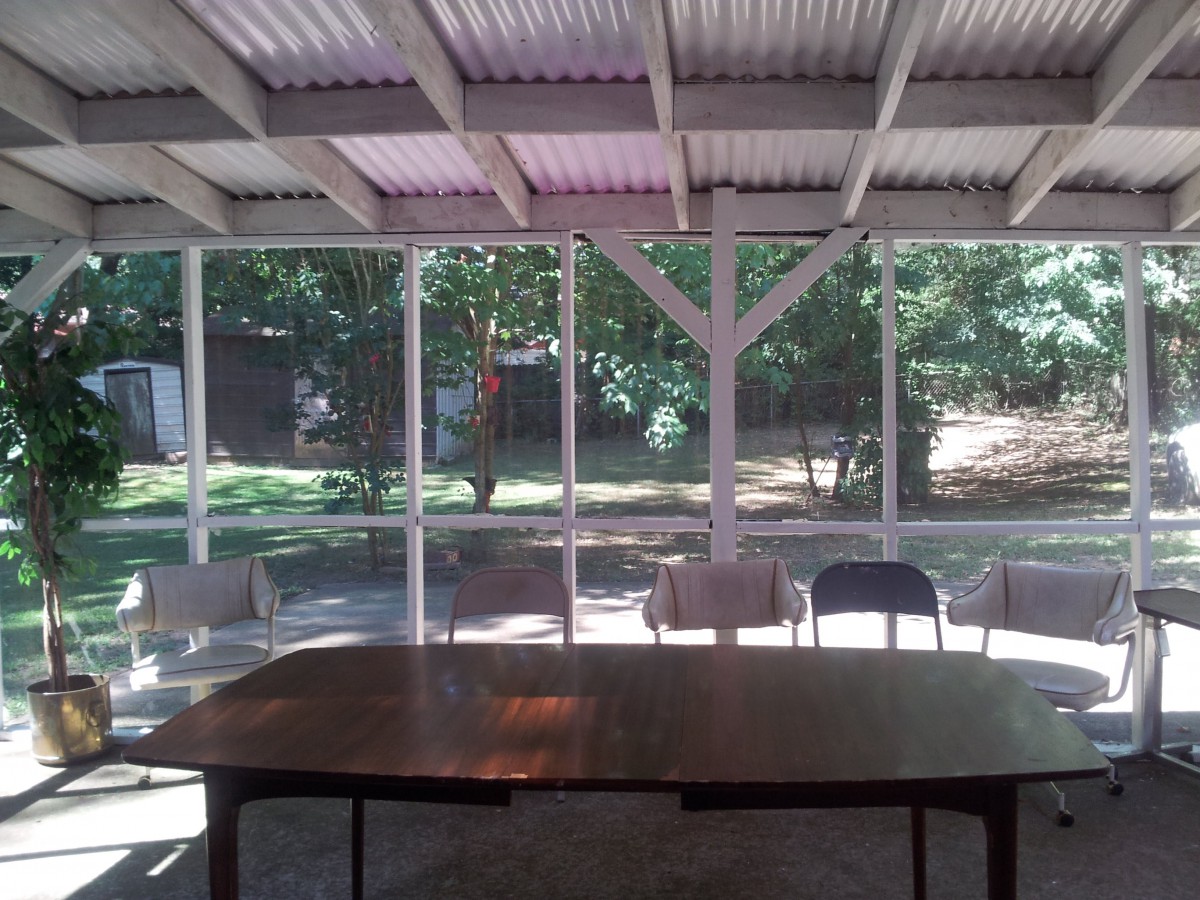 A covered outdoor area with a table and chairs, surrounded by trees and sunlight filtering through.