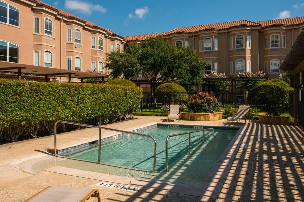 A well-maintained outdoor pool area surrounded by lush greenery and a large building under a clear blue sky.