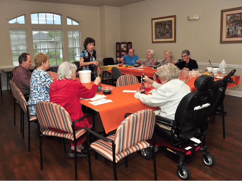 A group of seniors gathered around tables with orange tablecloths, engaging in a social meeting indoors.