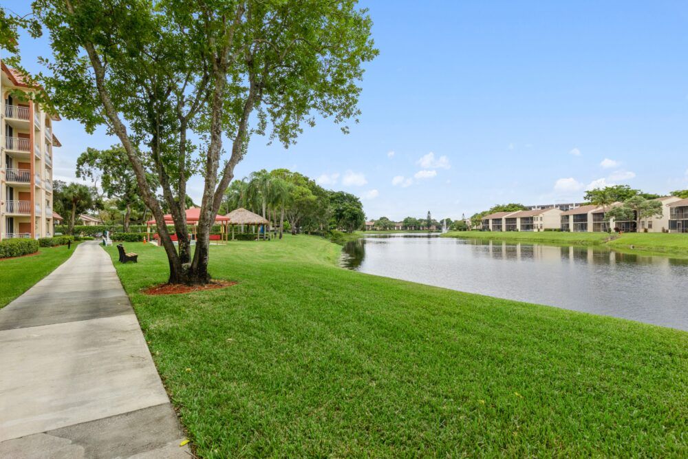 A serene residential landscape featuring a lakeside view with lush green grass, trees, and a pathway under a clear blue sky.