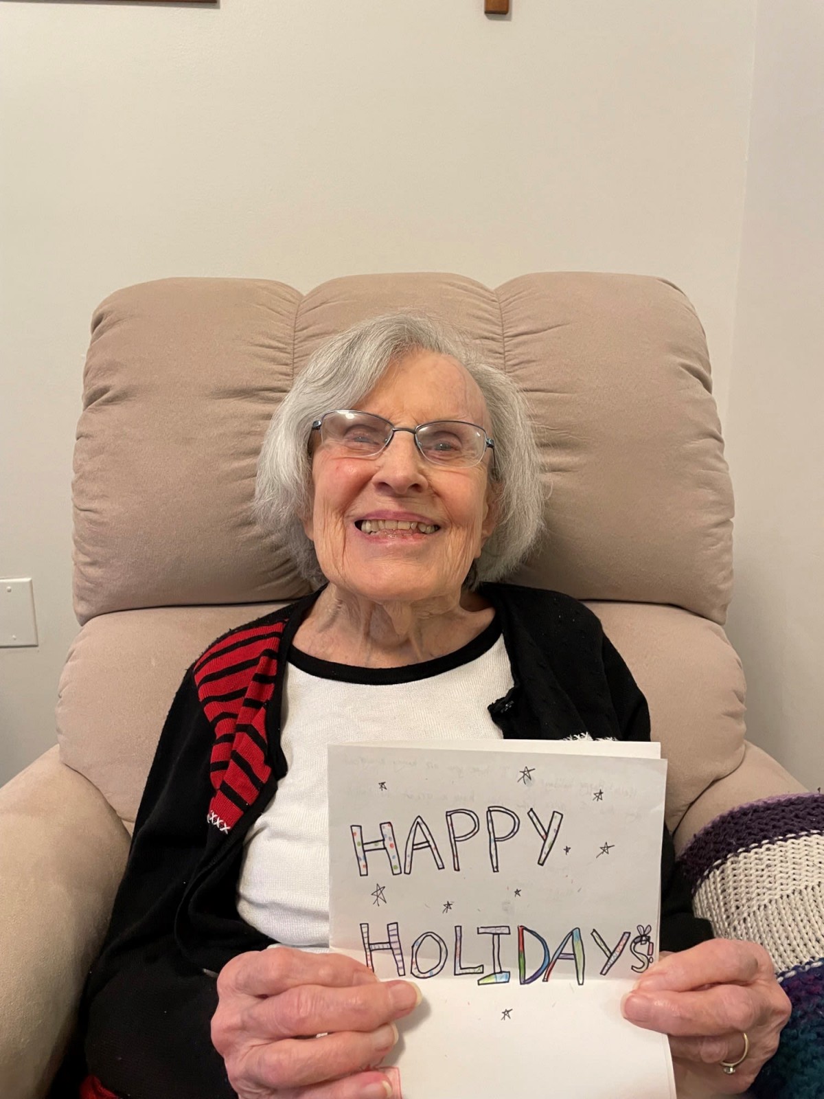 An elderly woman smiling while holding a 'Happy Holidays' sign indoors.