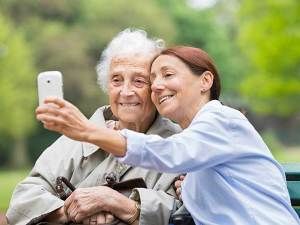 A younger woman is taking a selfie with an elderly woman outdoors, both smiling warmly.