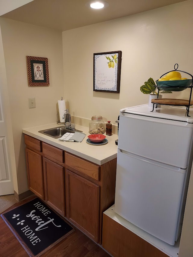 A compact kitchen area with a sink, countertop, and fridge, adorned with home decor and a fruit basket on top.