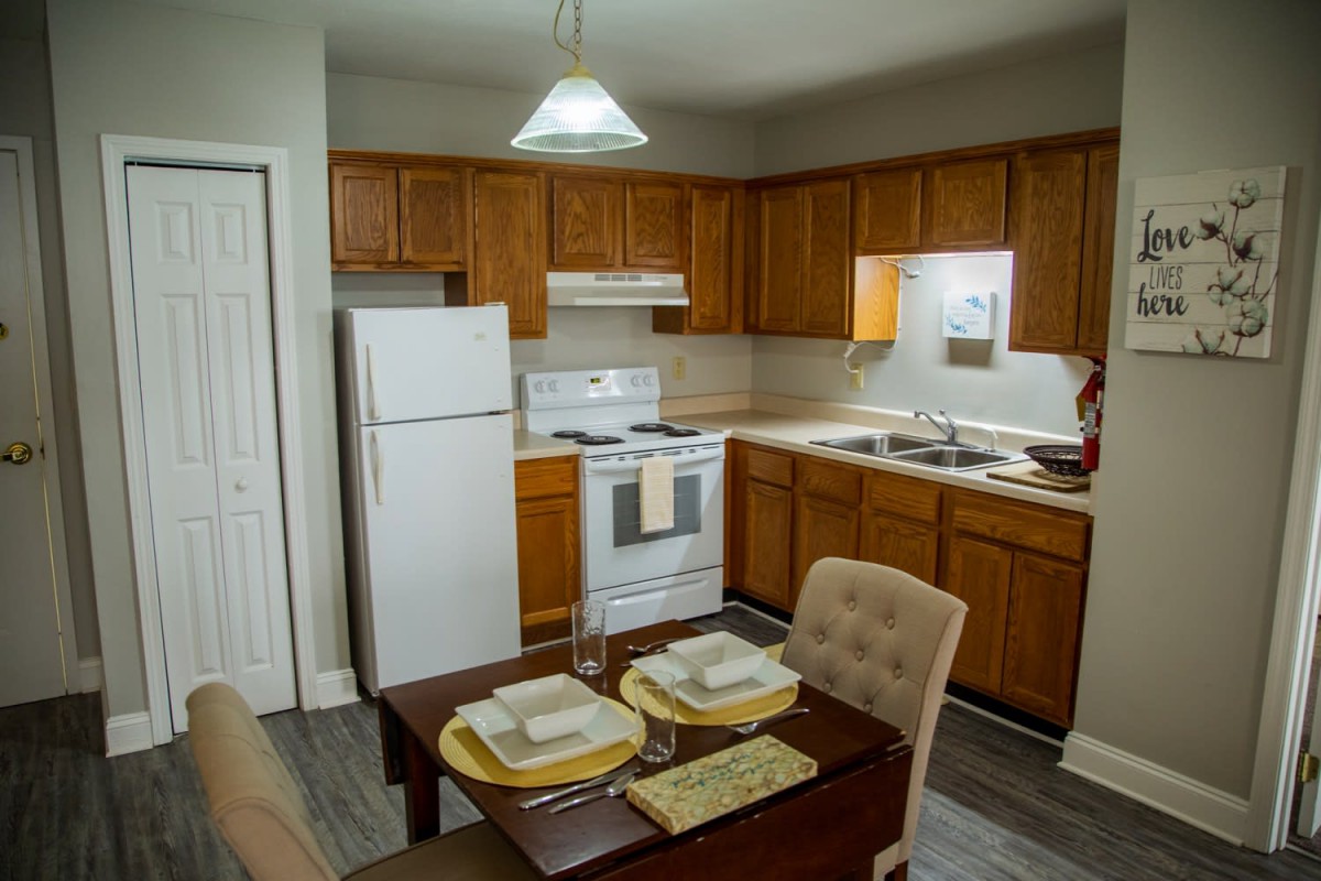 A cozy kitchen with wooden cabinets and a small dining area, featuring a refrigerator, stove, and a neatly set table.