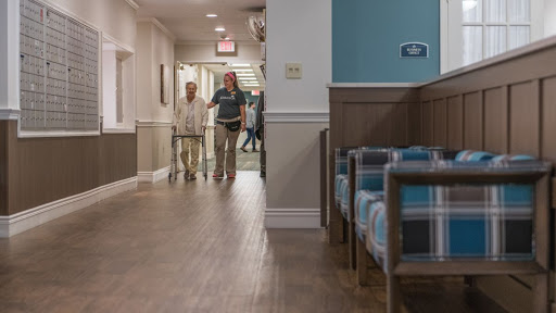 A caregiver assists an elderly person in a well-lit hallway of an assisted living facility.