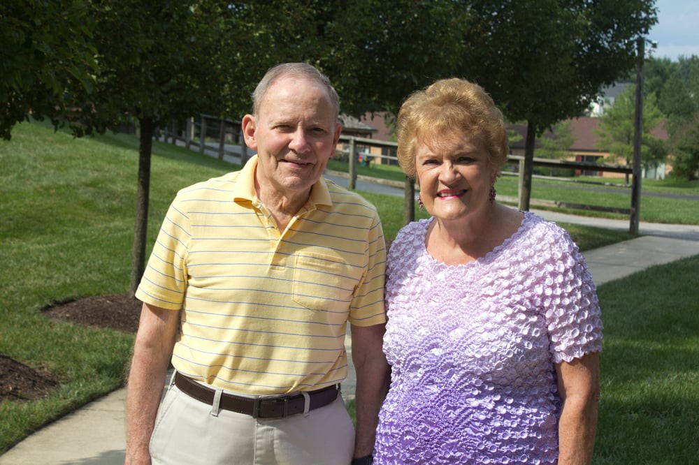 An elderly couple enjoying a sunny day outside, standing together with trees in the background.
