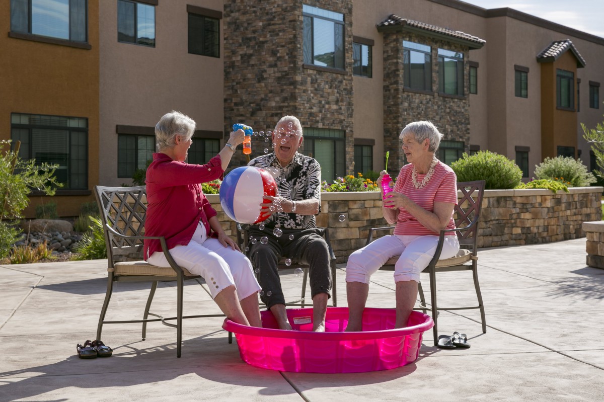 Three seniors enjoying a sunny day outdoors with their feet in a small pool, playing with a beach ball.