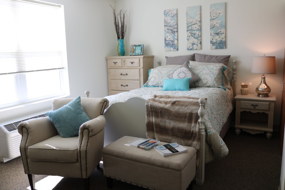 A cozy bedroom with elegant decor, featuring a bed, armchair, dresser, and wall art.