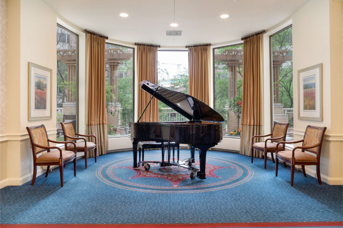 A grand piano in a bright music room with large windows and chairs.