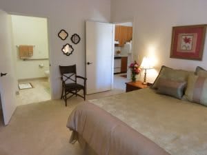 A cozy bedroom with neutral tones, featuring a bed, chair, lamp, and decorative elements.