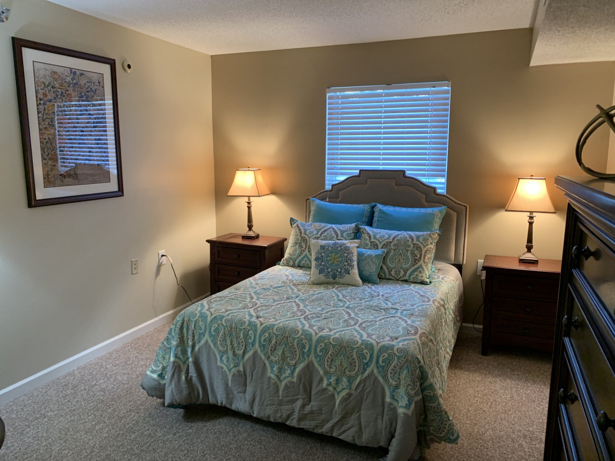A cozy bedroom with a neatly made bed between two wooden nightstands, each topped with a lamp, and a window providing natural light.