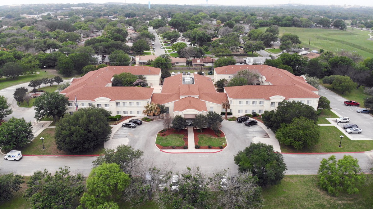 Aerial view of a large building complex with red roofs, surrounded by trees and a landscaped driveway in a suburban setting.