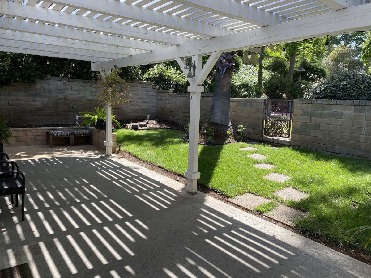 A sunny backyard with a pergola-covered patio and a small grassy area, surrounded by a fence.