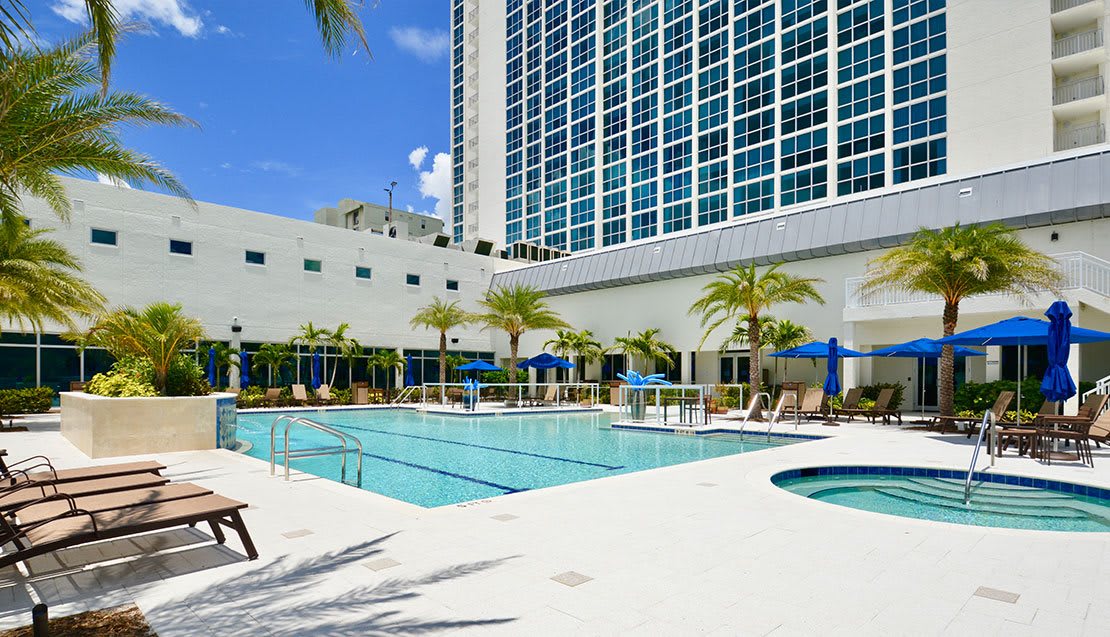 A modern hotel with a large outdoor swimming pool and palm trees on a sunny day.