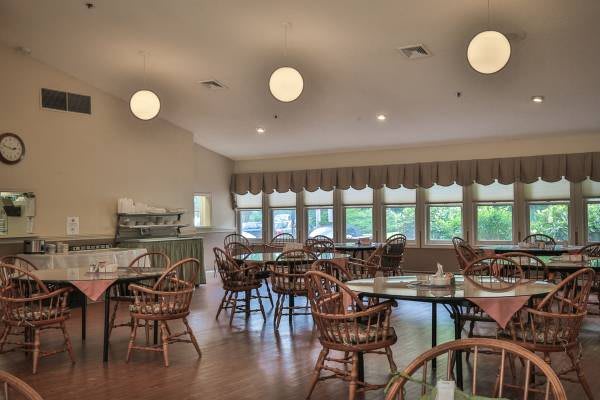 An empty dining room with wicker chairs and round tables, illuminated by ceiling lights and natural light from large windows.