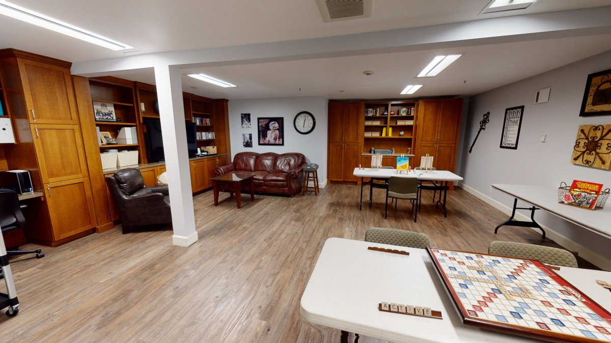 A cozy living room with wooden floors, a leather couch, bookshelves, and a game table featuring a Scrabble board.