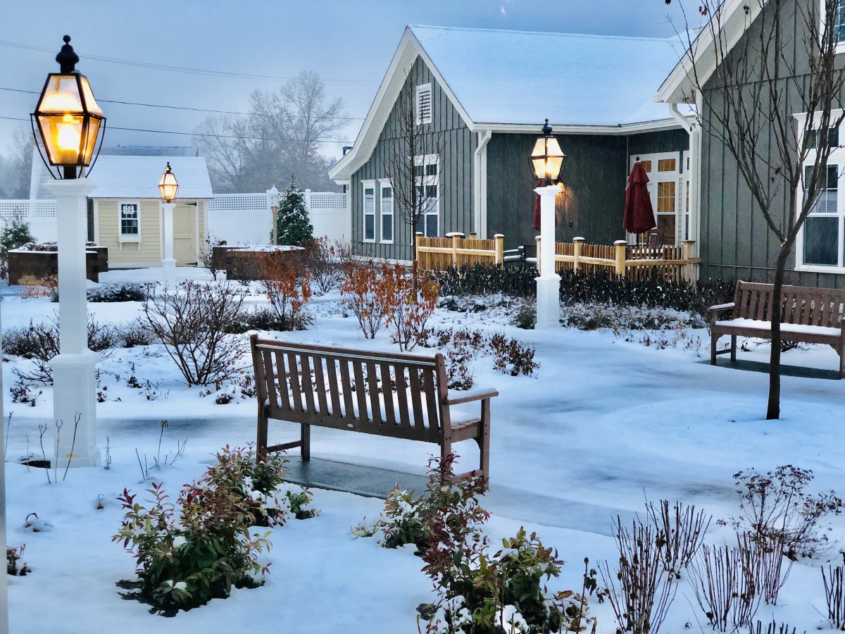 A snowy winter scene featuring a quaint house and garden with benches and a lamp post.