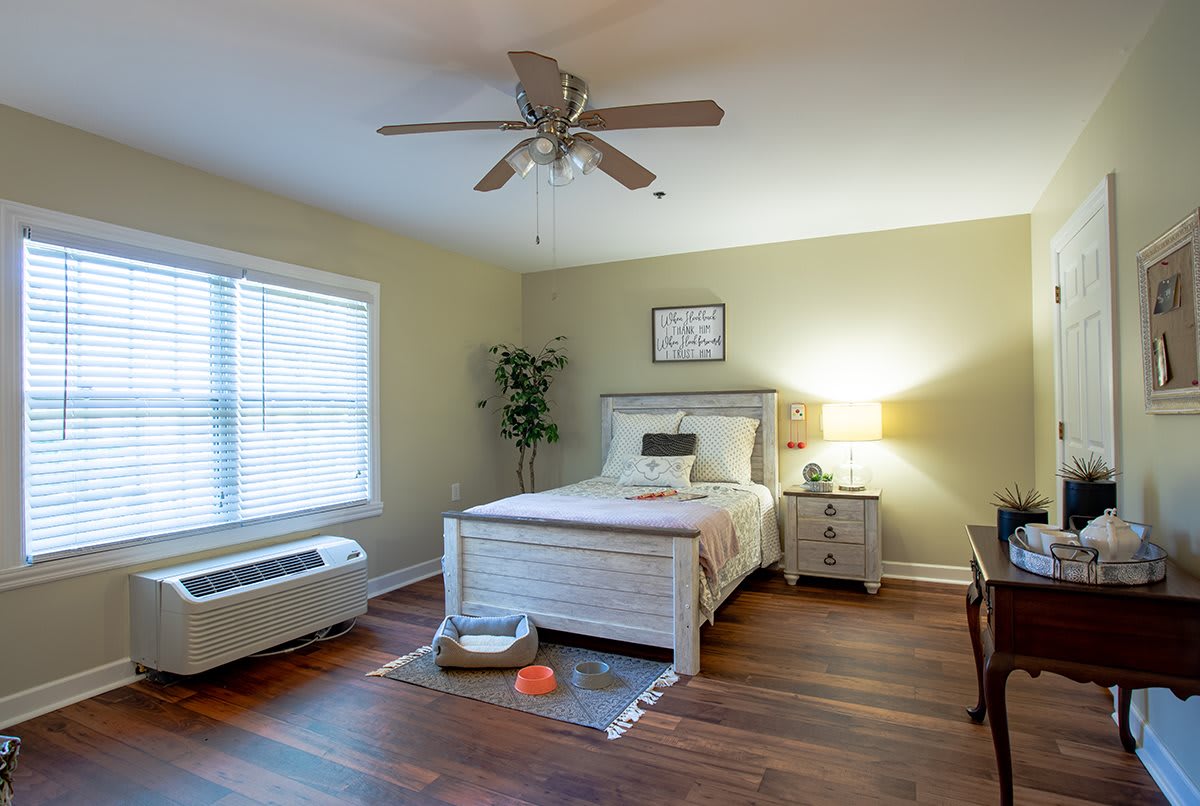 A cozy bedroom with a bed, nightstand, and ceiling fan, featuring hardwood floors and natural light from a window.