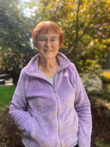 A woman wearing a purple jacket smiles while standing outdoors surrounded by greenery.