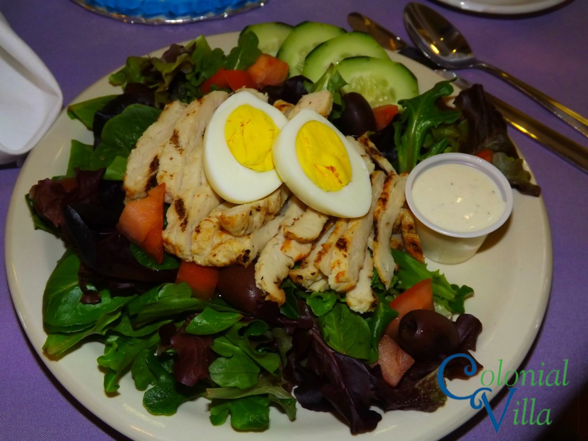 A fresh salad with grilled chicken, sliced hard-boiled eggs, cucumber, and tomato over mixed greens, served with a side of dressing.