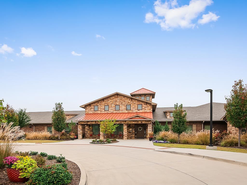 A large brick building with a driveway, surrounded by trees and bushes under a clear blue sky.