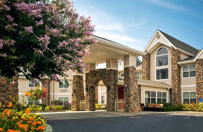 A picturesque building entrance with stone pillars, surrounded by trees and flowers under a clear sky.