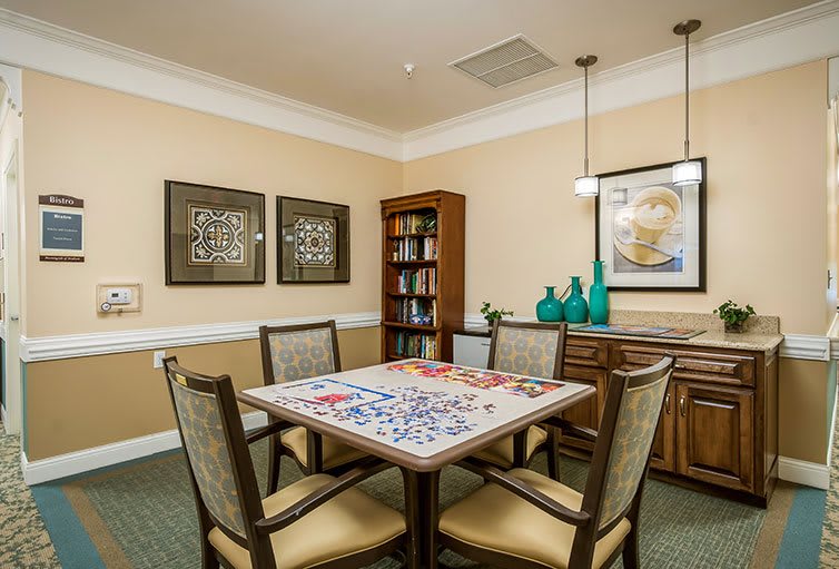 A cozy dining room with a table set for a puzzle, surrounded by chairs and decorated with artwork and vases.
