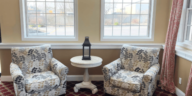 A cozy living room corner with two patterned chairs flanking a round table topped by a lantern, set against large windows.