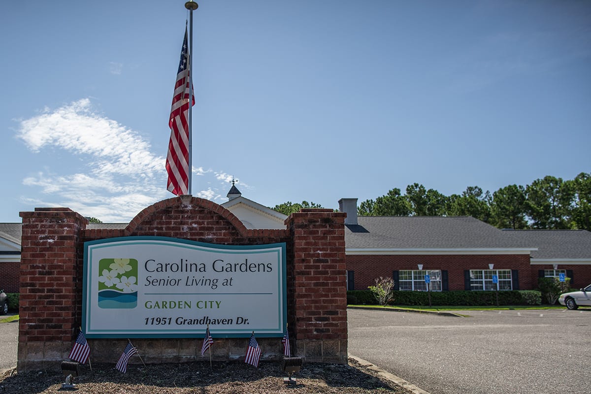 A brick building with an American flag and a sign for Carolina Gardens Senior Living at Garden City.