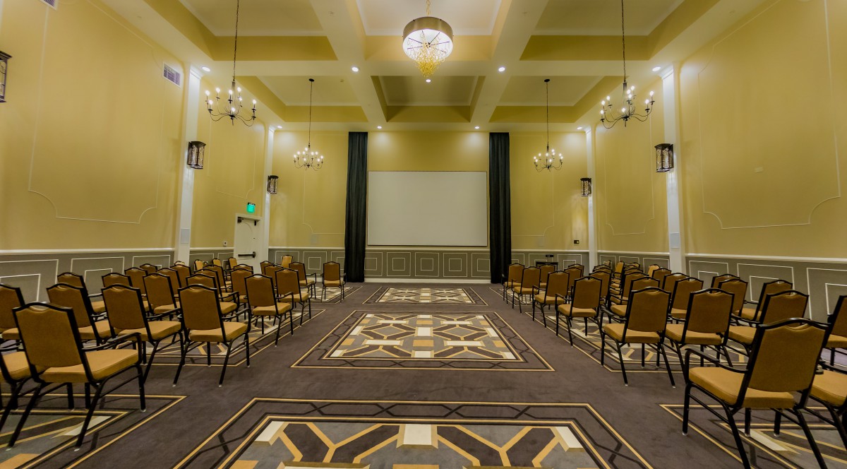 An elegant conference room setup with rows of chairs facing a large projector screen, illuminated by chandeliers.