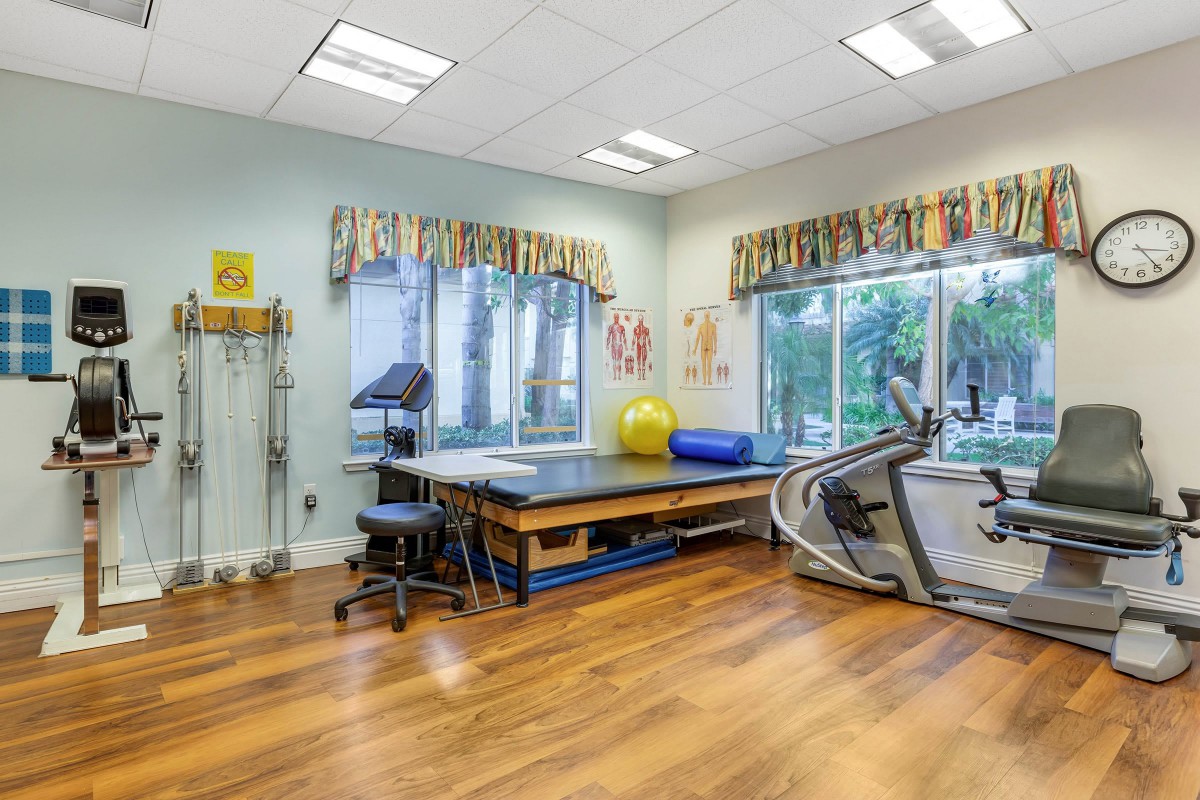 A well-equipped physical therapy room with exercise equipment and a treatment table, illuminated by natural light from large windows.