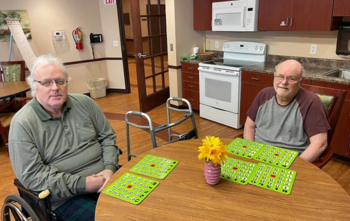 Two people sitting at a table in a kitchen area with bingo cards and a vase of flowers.