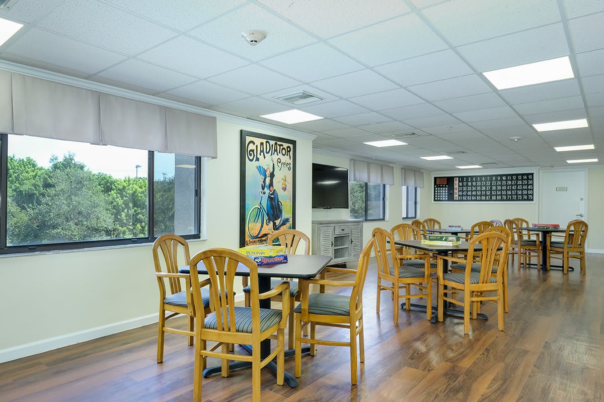 A bright community dining area with several tables and chairs, featuring large windows.