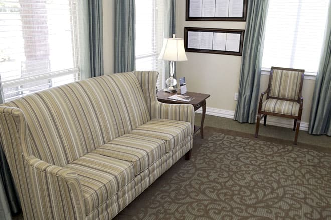A cozy living room featuring a striped sofa, a table with a lamp, patterned carpet, and elegant curtains.