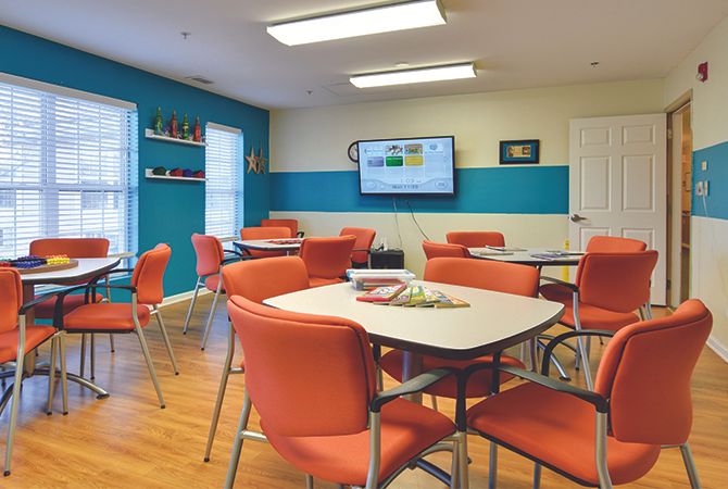 A modern classroom with orange chairs and tables, featuring a television on the wall.