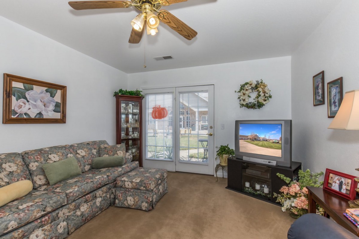 A cozy living room with floral-patterned sofa, ceiling fan, and a TV near a glass cabinet.