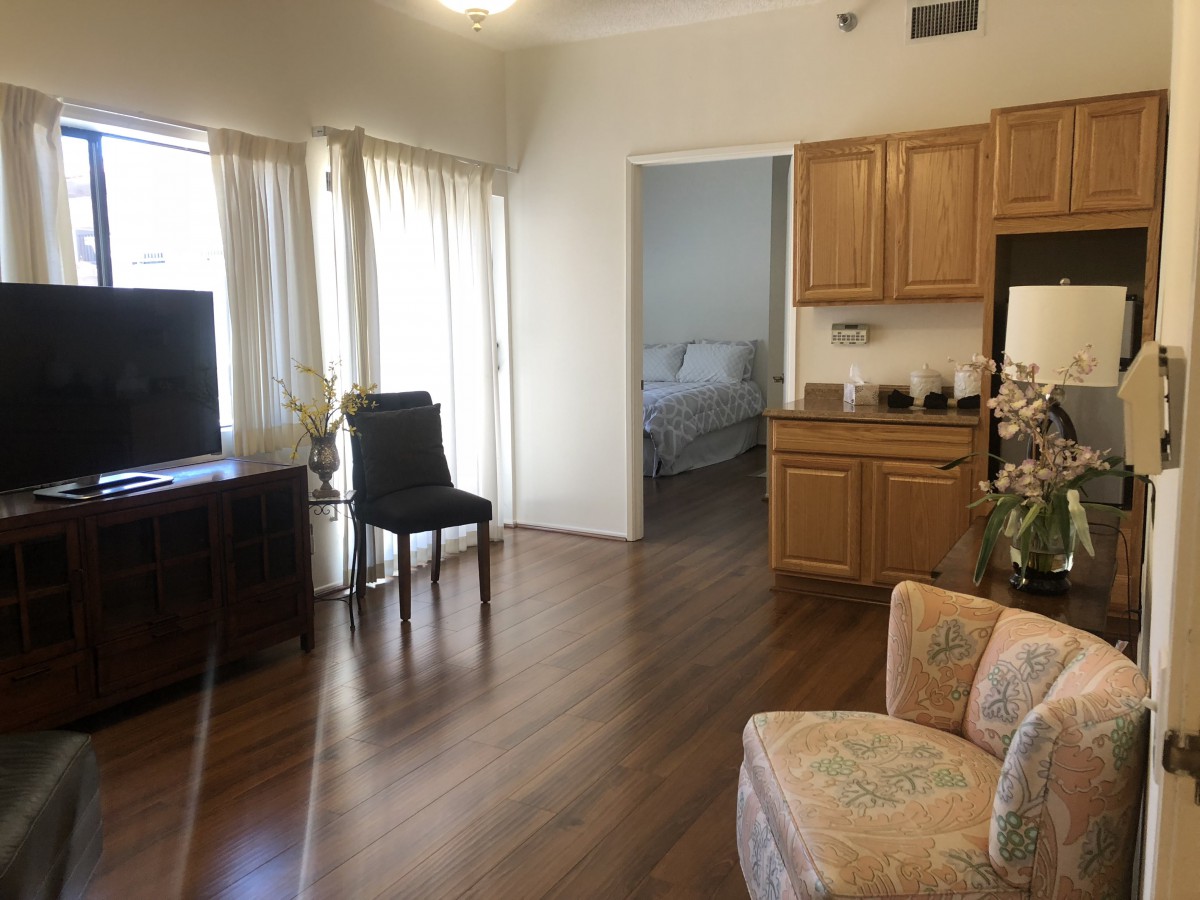 A bright living space with hardwood floors, featuring a TV, chairs, and kitchen cabinets.