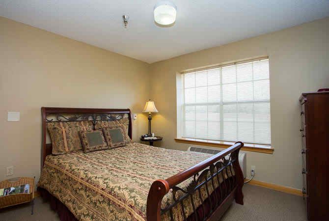 A cozy bedroom with a wooden bed, patterned bedding, a bedside lamp, and a window allowing natural light.