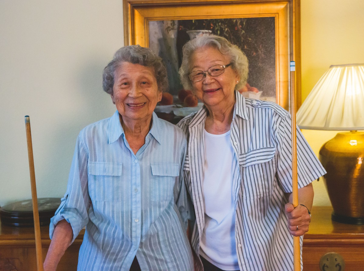 Two elderly women smiling and standing together indoors, both wearing striped shirts.