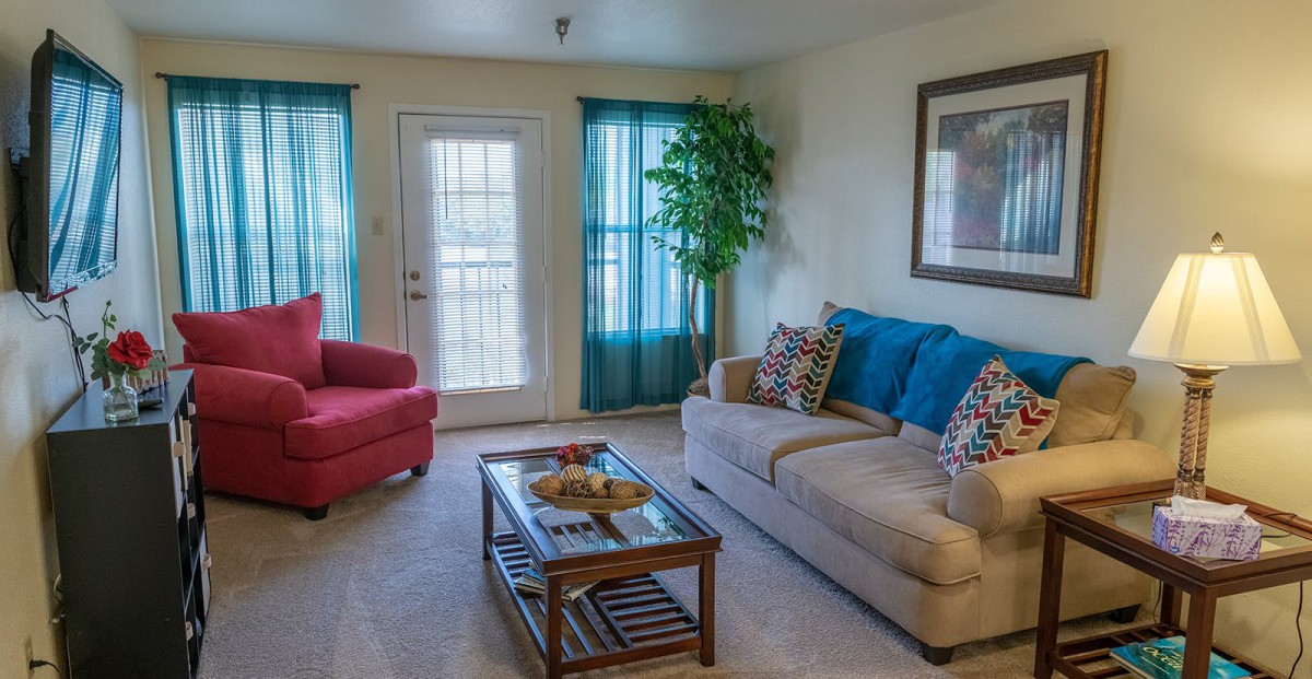 A cozy living room with a beige sofa and blue accents, a red armchair, a glass coffee table with decor, and bright natural light from the windows.