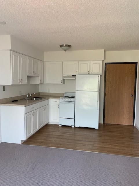 An empty apartment kitchen with white cabinets, a fridge, stove, and wooden flooring.