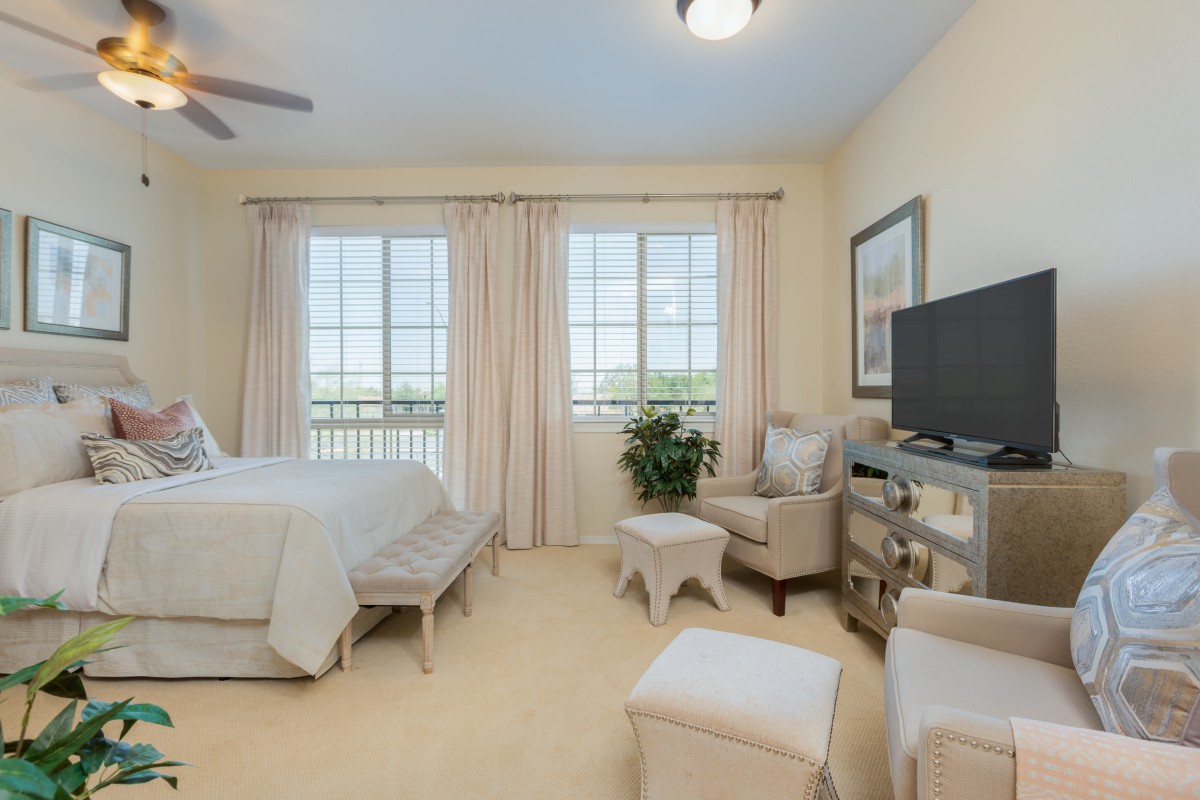 A well-lit bedroom with neutral tones, featuring a bed, armchair, television, and large windows with curtains.