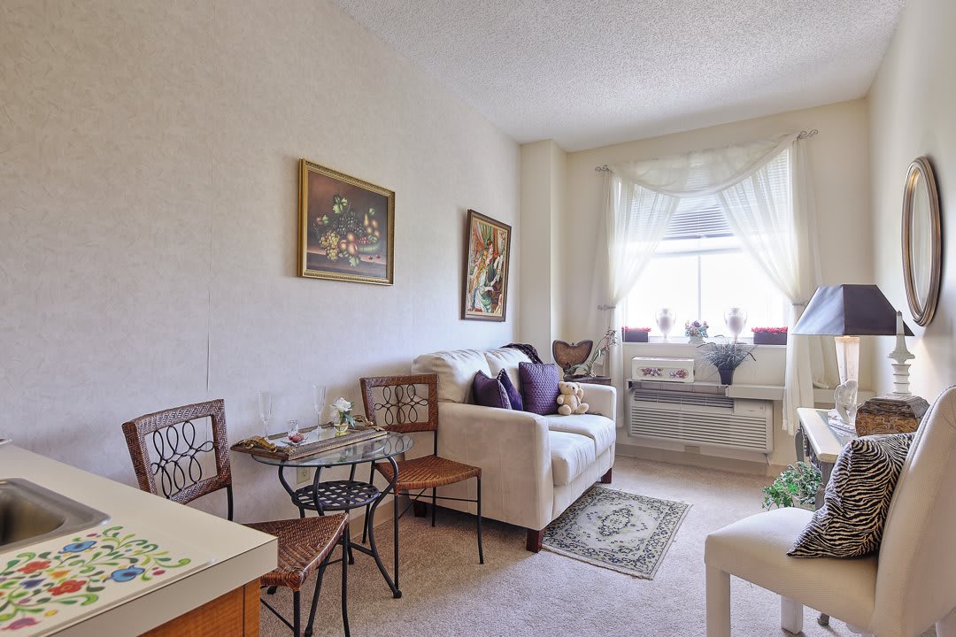 A cozy living room with a sofa, small dining table, artwork on the walls, and a sunlit window.