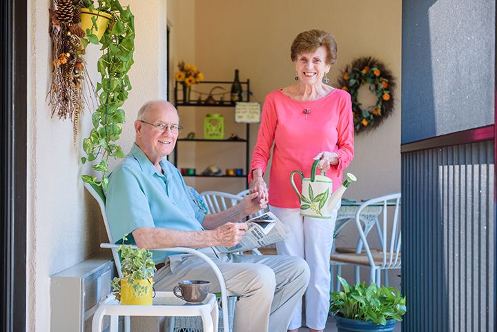 An elderly couple enjoys a sunny day on their balcony, tending to their home garden.