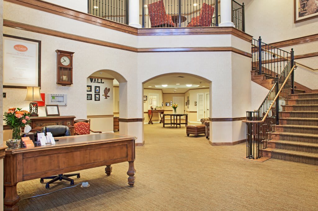 A spacious and elegant office interior featuring a wooden desk, a grand staircase, and classic decor.