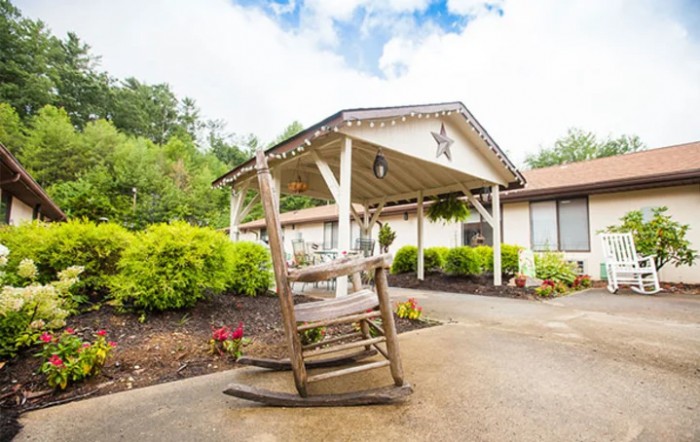 A rustic wooden rocking chair sits on a porch in front of a building with a landscaped garden and covered entrance.