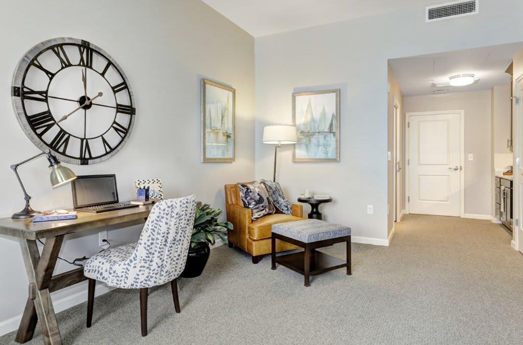 A stylish living room with a home office setup, featuring a large wall clock, artwork, and comfortable seating.