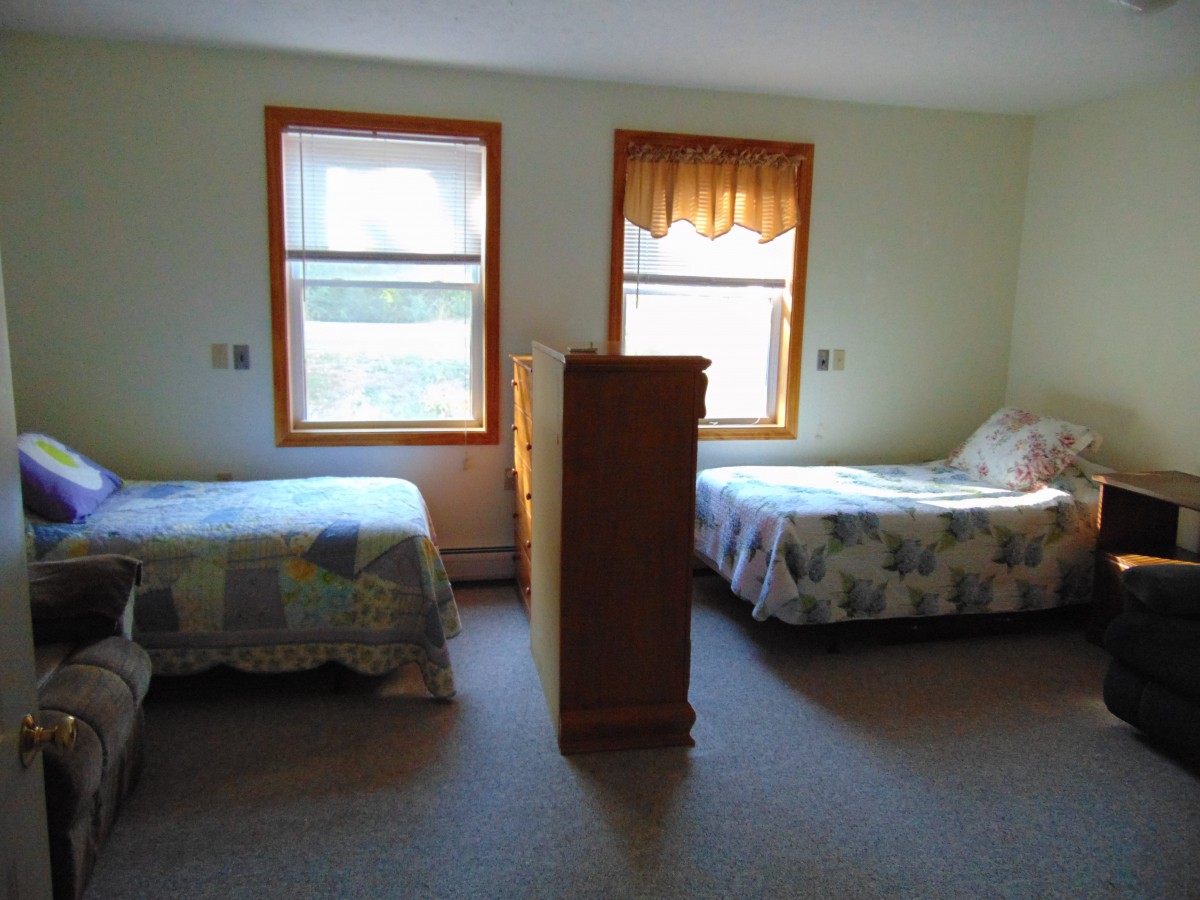 A simple bedroom with two beds separated by a dresser and sunlight streaming through the windows.