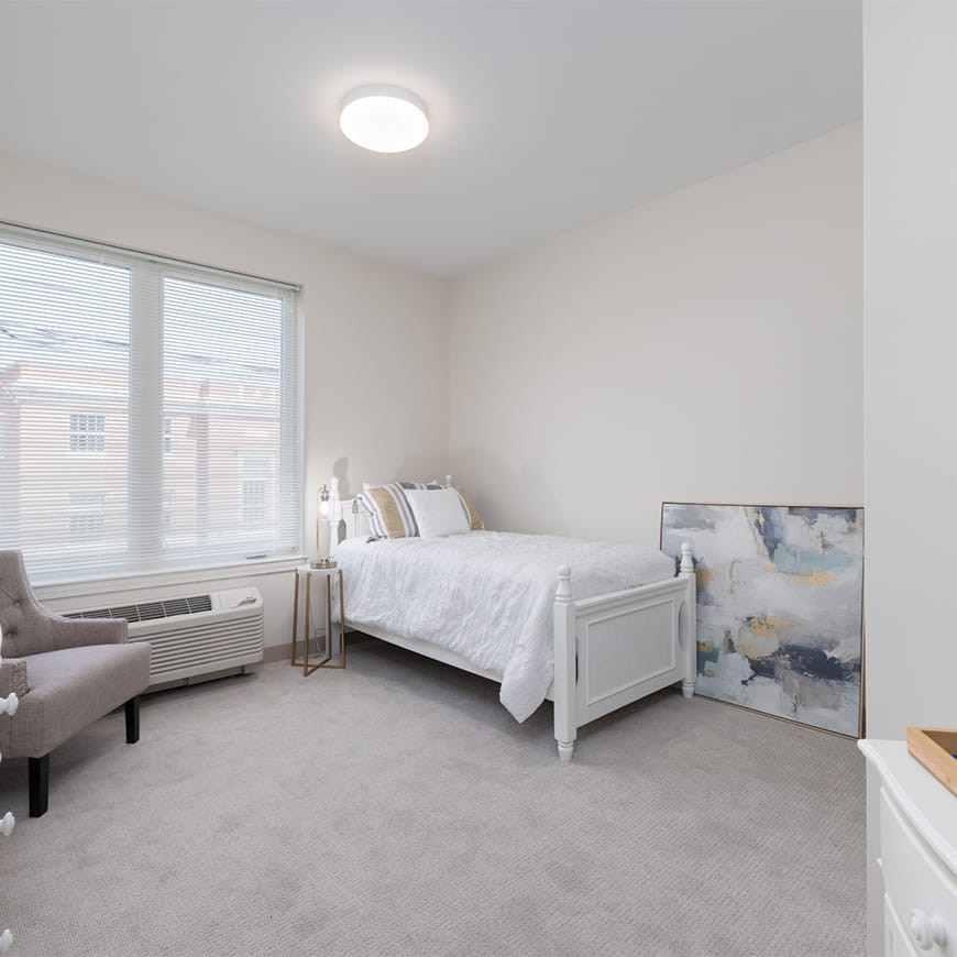 A minimalist bedroom with a single bed and soft neutral tones, featuring an armchair and wall art.